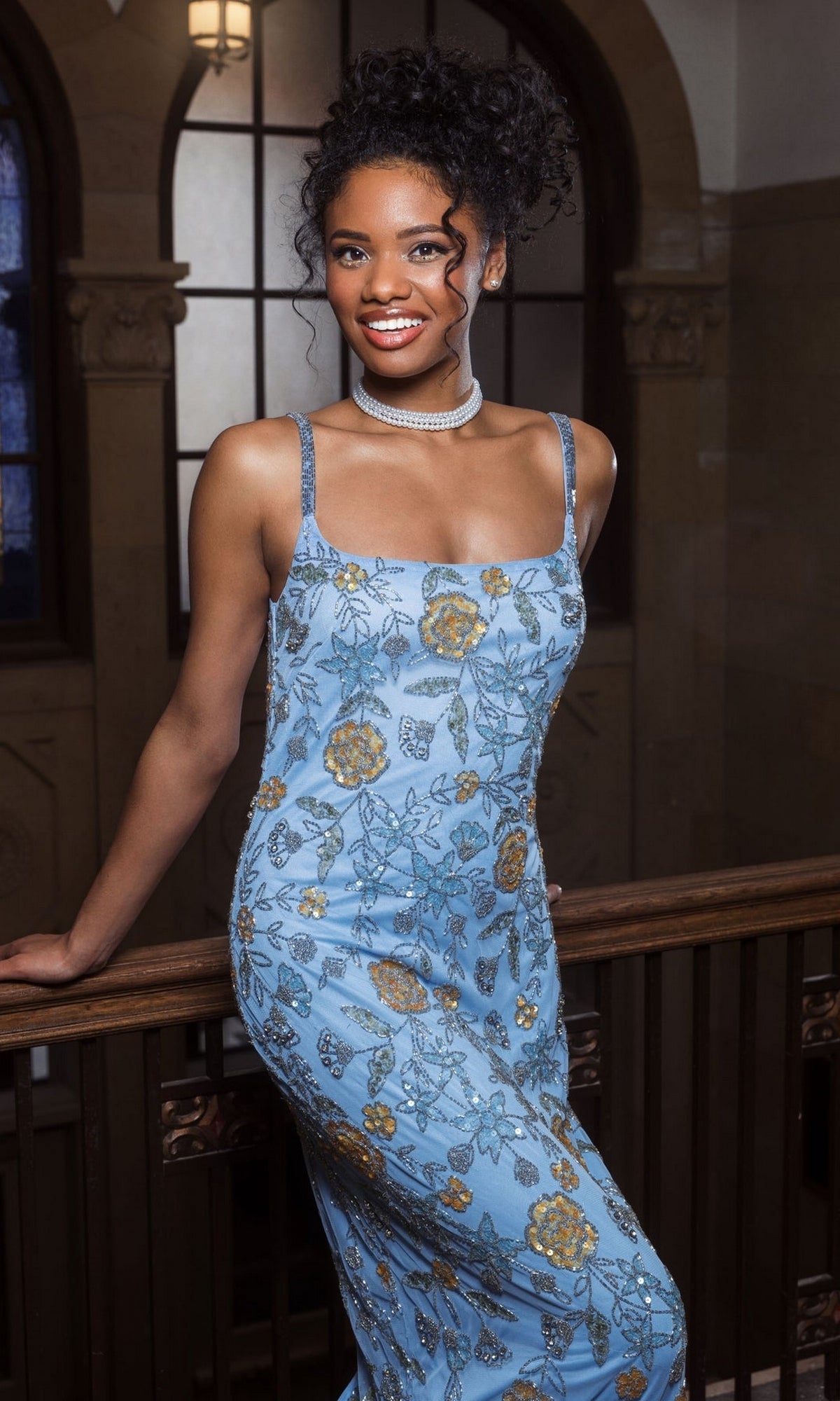 A woman wearing the Long Formal Dress 10808 by Mac Duggal, a blue floral beaded fitted tank dress, and a pearl necklace smiles at the camera while standing by a wooden railing indoors.
